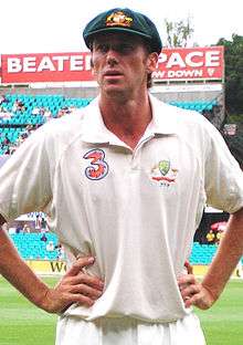 A white cricketer in cricket whites, wearing a baggy green cap. He has his hands on his hips and he is looking to his right.  He is standing in front of a bleacher.
