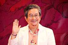 A man in front of a red curtain wears glasses and holds his right hand up.