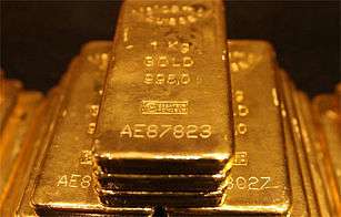 A hand holding a gold bar surrounded by several gold bars and other gold pieces in the background.