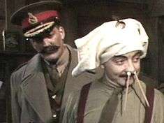 A man with a large moustache wearing a general's cap stands behind a man wearing underpants on his head and with pencils in his nose.