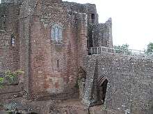 A castle, with a flat fronted tower facing the viewer with a stained glass window in the middle of it; a stone causeway is on the right of the picture, leading to a gateway to the right of the tower – a partially filled arch is supporting the causeway.
