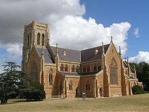 Exterior of Goulburn Cathedral. Built in honey-coloured stone, it has very large ornately-traceried windows in both the chancel and the transepts. Paired clerestory windows rise above the side aisles. The terminals of the gables have tall pinnacles. The tower which rises behind the chancel is square at the top.