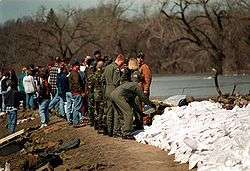 A line of people facing each other in an alternating manner are located next to a river. At the end of the line is a man putting a sandbag on top of a dike of numerous other sandbags. Multiple trees can be seen in the background.