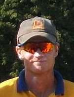 A man in a yellow shirt with a blue collar, wearing reflective sunglasses and a baseball cap, in front of foliage