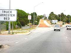 View of gavel-filled lane leaving the side of the road and then rising up a slope. Signs either side of it read 'TRUCK ARRESTOR BED'.