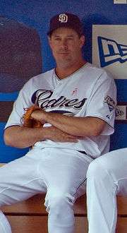 A man sits on a bench wearing a navy blue cap and a white baseball uniform in front of a blue wall