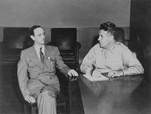 A large man in uniform and a bespectacled thin man in a suit and tie sit at a desk.
