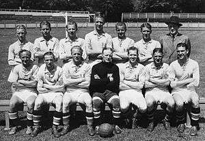 An association football team poses for a formative black-and-white photograph. A row of seven men sits on a bench, all wearing light-coloured shirts and white shorts apart from the player in the centre, who wears black. A football rests on the ground between his feet. Behind the seated row stand seven more men, all but one of whom are wearing the same light-coloured shirts. The exception is a gentleman standing at the end of the row on the viewer's right, who wears a dark double-breasted suit, tie and wide-brimmed fedora hat. All of those present have their arms folded apart from the man in the suit, whose hands are behind his back. In the background a set of goalposts can be seen.