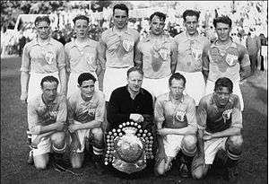 An association football team poses for a formative black-and-white photograph. A row of five men are kneeling on the pitch, all wearing light-coloured shirts and white shorts apart from the player in the centre, who wears black. In front of him is a large trophy shield. Behind the kneeling row stand six more men, all wearing the same light-coloured shirts. The players are noticeably tired; large sweat stains are visible on their shirts. A stand of a stadium can be seen in the background.