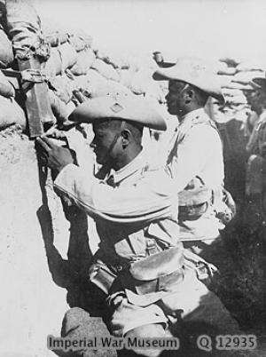 Two Gurkha soldiers in full uniform with turned-up slouch hats sit on the fire step in a fighting trench topped with sandbags. One soldier is looking through a periscope.