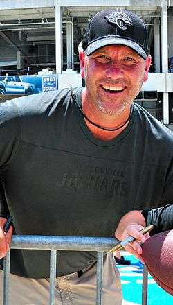 Color photograph of a smiling white man with salt-and-pepper goatee (Gus Bradley), wearing a dark grey Jacksonville Jaguars t-shirt and a dark teal Jaguars baseball cap.