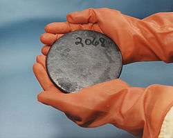 Two hands in brown gloves holding a blotched gray disk with a number 2068 hand-written on it