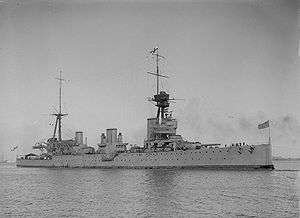 Side view of a large warship with three funnels and two large masts at rest on flat water.