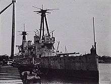 A World War I-era warship tied up to a dock. The ship is in poor condition, and has streaks of rust on her hull.
