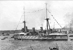 A World War I-era warship with tents erected over the fore and aft decks and flying flags from her rigging. Other warships, a small fortress and land are visible behind the ship and several small boats are visible in the foreground.
