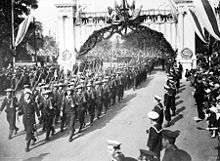 A group of men wearing military uniforms and carrying rifles marching under a decorated arch. The group is being led by a man carrying a sword. They are being watched by other men in uniform and a crowd of civilians.