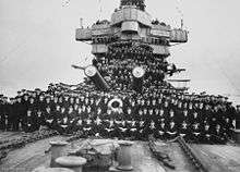 A large number of men posing for a photo on the foredeck of a warship. Two of the ship's gun barrels are visible in the middle of the group.