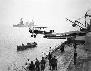 A small biplane has just taken off from a warship, having used a platform built over the roof and barrels of a twin-barrelled turret. Sailors are observing the launch from the deck of the ship, and from a small boat nearby. A large warship is visible in the background.