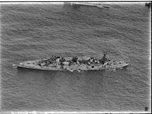 An aerial photograph of a large, World War I-era warship armed with four gun turrets, each with two guns, at sea. The ship is listing to port, but does not appear to be otherwise moving.