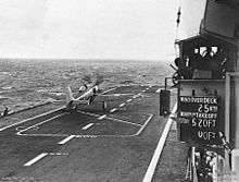 The deck of an aircraft carrier with a propeller aircraft towards the front of the ship. Some of the ship's superstructure is at right, including a board displaying the wind conditions