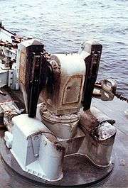 An empty missile launcher on the front of a warship, it has scorch marks on it. In the background there are empty shell casings.