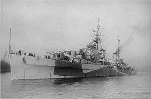 Black and white photo of a World War II-era warship. The ship is painted in camouflage and is armed with several gun turrets.