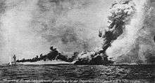 A large warship is almost completely obscured by a huge column of smoke.