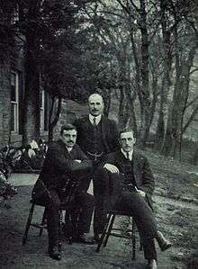 Three men in dark suits, two sitting on chairs and the third stood behind
