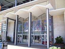 glass lobby with several columns in front holding a roof to shelter the doorway, with a tile mosaic of sports figures inside the lobby
