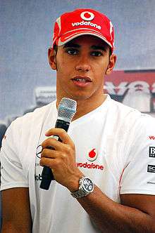 A man in his early twenties wearing a white T-shirt and a red baseball cap with sponsors logos. He is holding a microphone in his left hand.