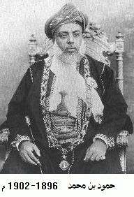 A black-and-white photograph of a man with a white beard wearing a turban, a dark jacket, a white shirt, and a belt and sitting on a chair