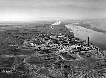 A cluster of industrial structures lie on a flat plain by a big river. The structures include a few low rectangular buildings, many smaller buildings, cylindrical tanks of varied sizes, and a tall smokestack. Several roads connect the cluster to other parts of the plain. Smoke or steam rises from two places further upriver.