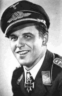 The head and shoulders of a young smiling man, shown in semi-profile. He wears a field cap and a military uniform, with an Iron Cross displayed at the front of his shirt collar.