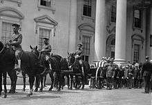 A black-and-white photograph of a procession of people in uniforms, some of whom are riding horses, all standing in front of a white building