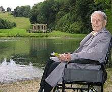An elderly man in a wheelchair near a small lake in the English countryside.