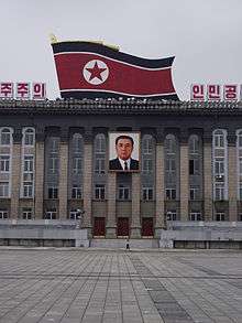 Large, columned building, with black, red and white flag and portrait of leader