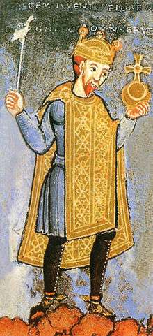 A mediæval illustration of a man with a short red beard wearing a blue tunic and a gold over-tunic, with black tights, holding a golden orb in his left hand and a silver sceptre in his right. Above his red hair, he is wearing a gold crown. Indistinct words are faintly visible above him.