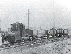 The driver stands beside a small electric locomotive towing four coal wagons