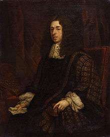 A dour-looking man sitting in a simple, studded, leather chair. He is wearing an ornate black robe with gold trim, and possibly a large brown wig, although that may be his hair. In his hand is a piece of paper.