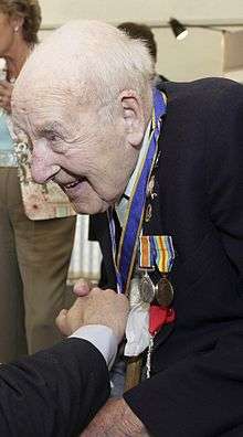 An elderly man, leaning forward as he shakes someone's hand, wearing several medals