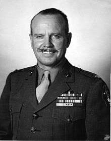 Head & torso of white man with slightly receding hairline and waxed mustache wearing tan shirt and necktie, green blouse with U.S. Marine Lieutenant Colonel's insignia, five rows of ribbons on his chest, and 2nd Marine Division patch on his left shoulder. The U.S. Marine Corps logo is superimposed on the lower right corner of this black & white photograph.