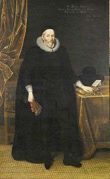 A tall elderly man with a beard, wearing long black robes and a large white ruff.  He is standing with a fan in his right hand and with his left hand resting on books on a table.