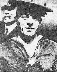 Head and shoulders of a white man wearing a flat sailor's cap, a sailor suit with scarf tied around the neck under the collar, and a star-shaped medal hanging from a ribbon around his neck.