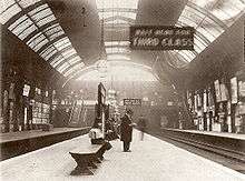 A black-and-white photograph of a railway station platform under a barrel roof. Several figures are visible, one standing wearing a top hat, a sign reads "WAIT HERE FOR THIRD CLASS".