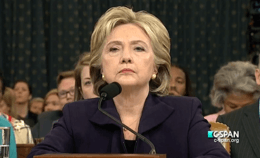 Clinton testifying before the House Select Committee on Benghazi on October 22, 2015. She is standing behind a podium with a microphone, with many people behind her.