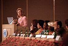 Clinton speaking at a podium with several onlookers. She is delivering her famous "human rights are women's rights and women's rights are human rights" speech in Beijing during September 1995.