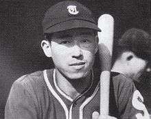 Hiroshi Ohshita wearing a dark old-style baseball uniform resting a baseball bat on his left shoulder