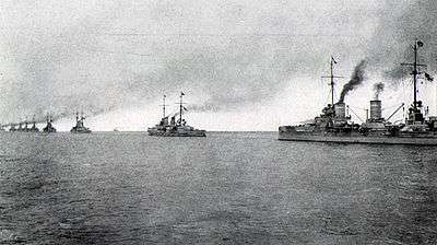 A line of nine large gray battleships stretches into the distance, all belching dark black smoke from their funnels