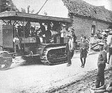 A Holt seventy-five tractor towing a field gun through a war-damaged village in Europe. The tractor is stacked high with supplies, and a number of uniformed soldiers are walking alongside.