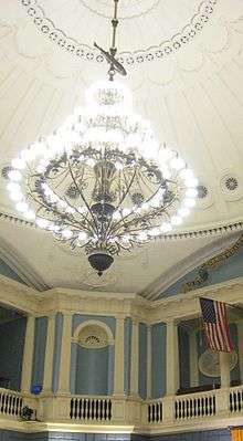  A room with high ornate blue-and-white walls and a white-domed ceiling, from which a large chandelier hangs by a rod. Along the rod, between the chandalier and the ceiling, is the figure of a fish.